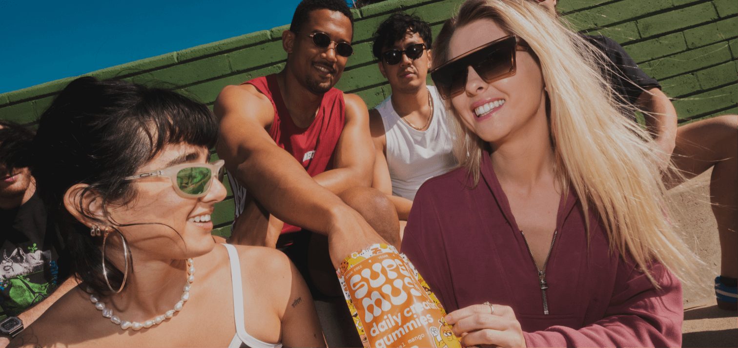 Friends on sunny bleachers passing a bright orange pouch of SuperMush gummies